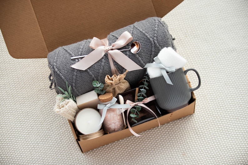 K&ouml;nnte beinhalten: Eine Geschenkbox mit Artikeln zur Selbstpflege. Sie enth&auml;lt eine graue Strickdecke mit rosa Schleife, eine graue Tasse, eine Kerze, Seife, Badesalz und einen kleinen Jutesack. Die Artikel sind in einer braunen Pappschachtel angeordnet.