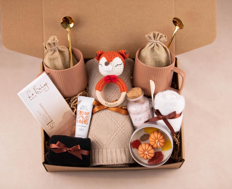 May include: A gift box containing pink mugs with gold spoons, a chocolate bar, hand cream, a black washcloth with a brown bow, a candle with pumpkin decorations, and a knitted fox rattle. The box also includes a knitted hat.