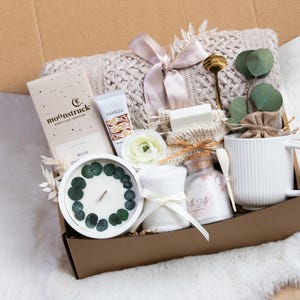 May include: A gift box filled with self-care items. Includes a white candle with eucalyptus, a mug, a bar of soap, a tube of vanilla cream, a chocolate bar, and a knitted blanket. The box is tied with a pink ribbon.