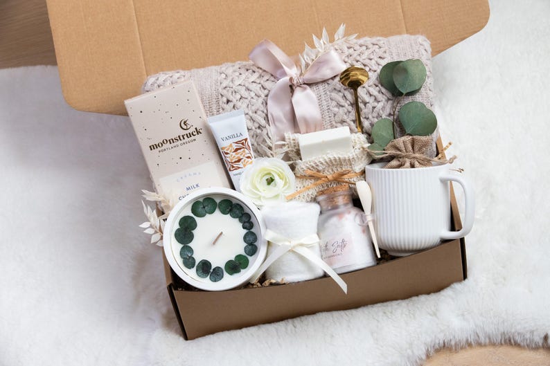 May include: A gift box filled with self-care items. Includes a white candle with eucalyptus, a mug, a bar of soap, a tube of vanilla cream, a chocolate bar, and a knitted blanket. The box is tied with a pink ribbon.