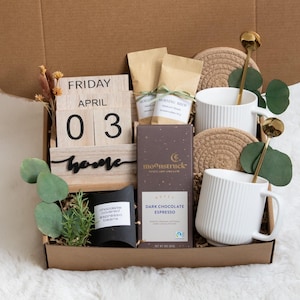 May include: A gift box filled with coffee, chocolate, and mugs. The box contains a wooden calendar displaying "Friday April 03" and the word "home". Two white mugs with gold spoons, coasters, and a dark chocolate bar are also included.