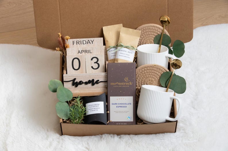 May include: A gift box filled with coffee, chocolate, and mugs. The box contains a wooden calendar displaying "Friday April 03" and the word "home". Two white mugs with gold spoons, coasters, and a dark chocolate bar are also included.