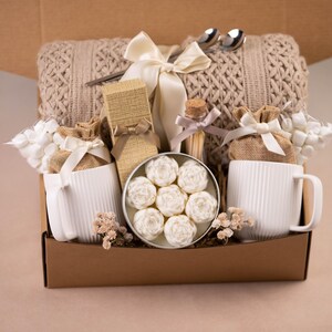 May include: A gift box filled with a beige knitted blanket, two white mugs, and a variety of small gifts. The box includes a tin of white floral-shaped soaps, a small glass bottle, and two silver spoons.