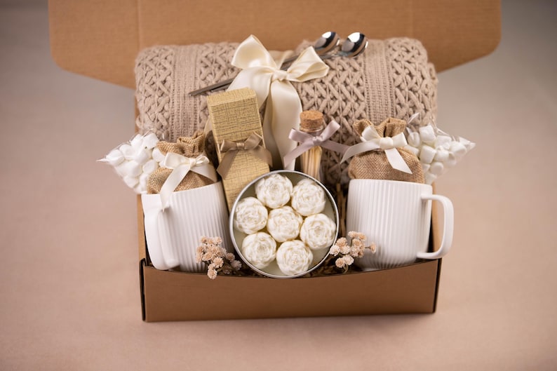 May include: A gift box filled with a beige knitted blanket, two white mugs, and a variety of small gifts. The box includes a tin of white floral-shaped soaps, a small glass bottle, and two silver spoons.