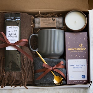 May include: A gift box filled with cosy items for a relaxing evening. It includes a brown scarf, a grey mug, a bag of coffee labelled "coffee early bird blend", a gold spoon, a dark chocolate espresso bar, a candle, and a small bag of sweets.