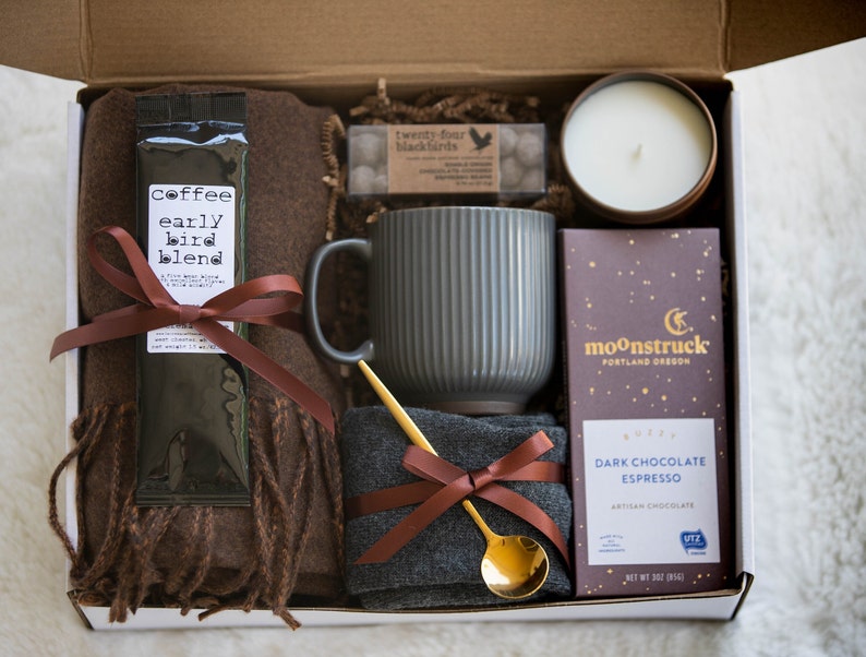 May include: A gift box filled with cosy items for a relaxing evening. It includes a brown scarf, a grey mug, a bag of coffee labelled "coffee early bird blend", a gold spoon, a dark chocolate espresso bar, a candle, and a small bag of sweets.