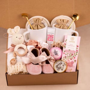 May include: A gift box filled with baby items and self-care products. Includes pink baby shoes, a crochet bunny rattle, white mugs, a bar of rose cardamom chocolate, and a bath bomb. The box is brown.