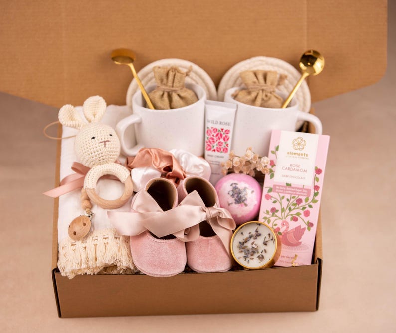 May include: A gift box filled with baby items and self-care products. Includes pink baby shoes, a crochet bunny rattle, white mugs, a bar of rose cardamom chocolate, and a bath bomb. The box is brown.