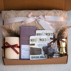 May include: A gift box filled with a cozy blanket, a candle, a chocolate bar, a cocktail mix, a martini glass, and a jigger. The box is decorated with a brown ribbon and a bow.