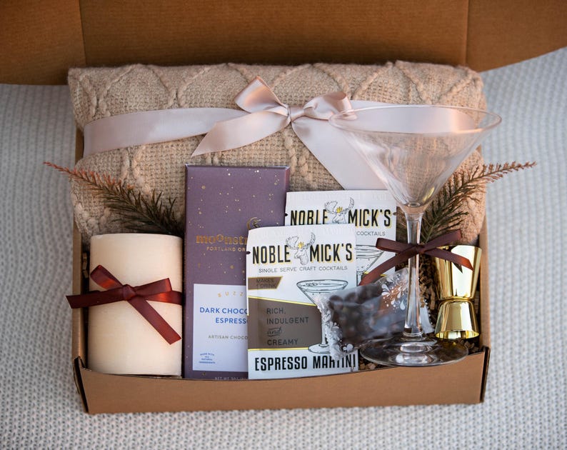 May include: A gift box filled with a cozy blanket, a candle, a chocolate bar, a cocktail mix, a martini glass, and a jigger. The box is decorated with a brown ribbon and a bow.