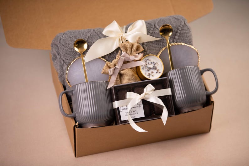 May include: A gift box filled with a gray knitted blanket, two gray ribbed mugs, gold spoons, coasters, a candle, and a box of dark chocolate caramels. The box is tied with a white ribbon.