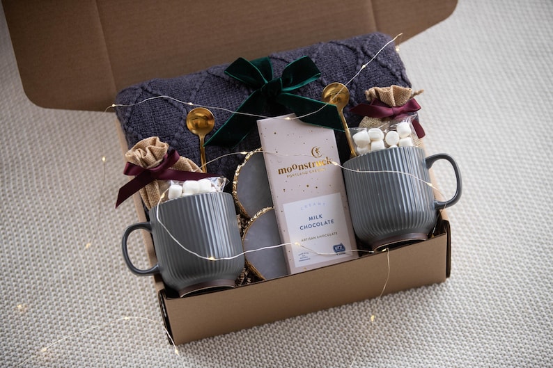 May include: A gift box filled with a gray knit blanket, two gray mugs, marshmallows, chocolate, and gold spoons. The box is tied with a green velvet bow and contains decorative string lights.