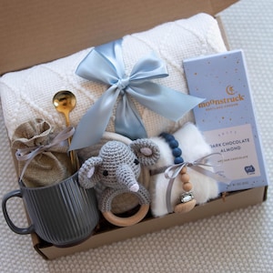 May include: A baby gift basket with a white blanket, a gray mug, a wooden rattle, a gray crocheted elephant, a small bag, a blue ribbon, a spoon, a chocolate bar with the text "moonstruck", and a wooden bead necklace.