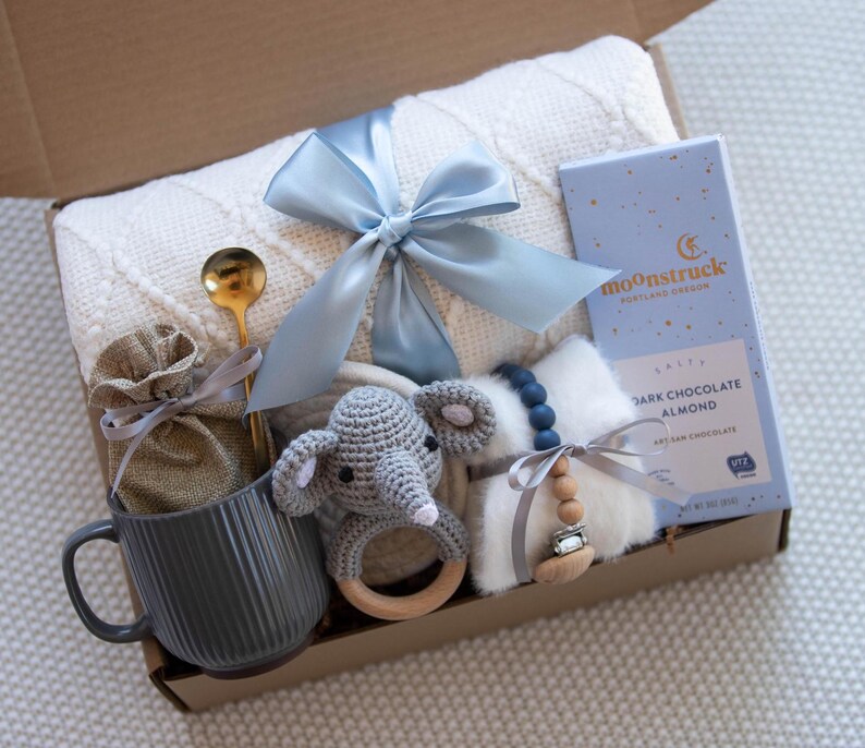 May include: A baby gift basket with a white blanket, a gray mug, a wooden rattle, a gray crocheted elephant, a small bag, a blue ribbon, a spoon, a chocolate bar with the text "moonstruck", and a wooden bead necklace.