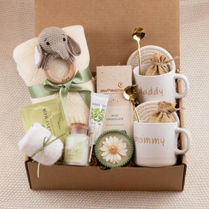 May include: A gift box filled with baby and self-care items. Includes a cream-colored blanket, a wooden elephant teether, two mugs that say "daddy" and "mommy", and other items like eye masks, a candle, and chocolate.
