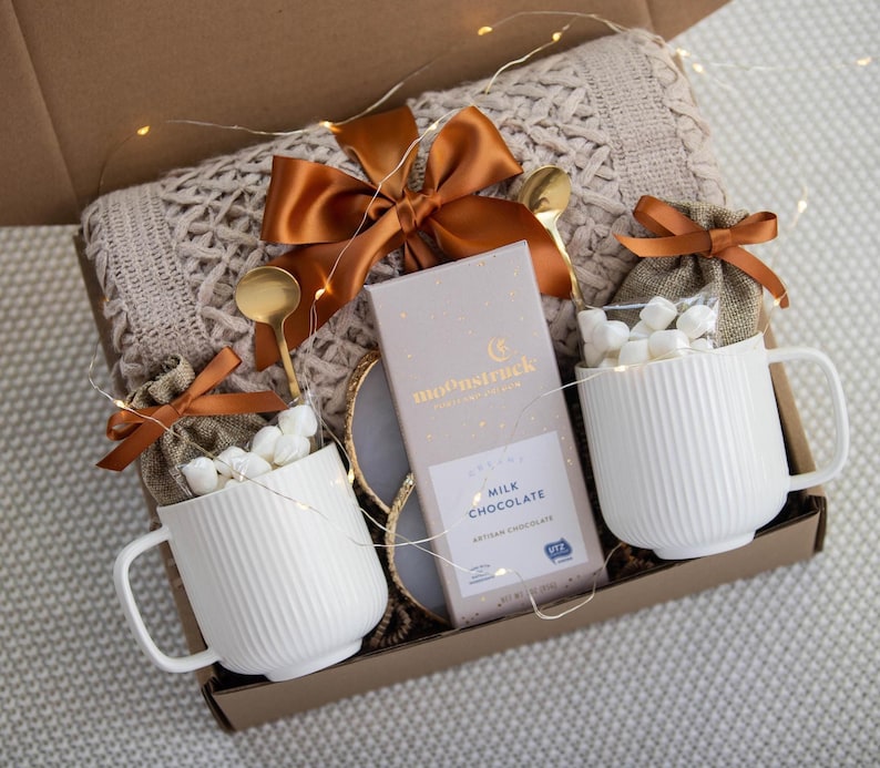 Puede incluir: Una caja de regalo con dos tazas blancas acanaladas llenas de malvaviscos, cucharas doradas y una barra de chocolate con leche "Moonstruck". Una manta de punto trenzado beige y un lazo naranja completan la presentaci&oacute;n. Las luces de hadas a&ntilde;aden un toque festivo.