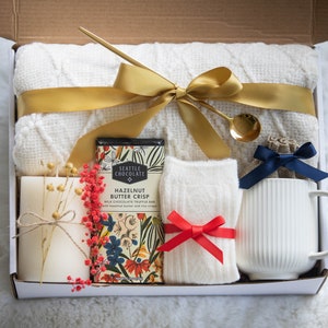 K&ouml;nnte beinhalten: Geschenkbox gef&uuml;llt mit einer wei&szlig;en Decke, einem goldenen Band, einer Schokoladenriegel, einer Kerze, Socken mit roter Schleife, einer Tasse und einem kleinen goldenen L&ouml;ffel. Die Box ist ge&ouml;ffnet.