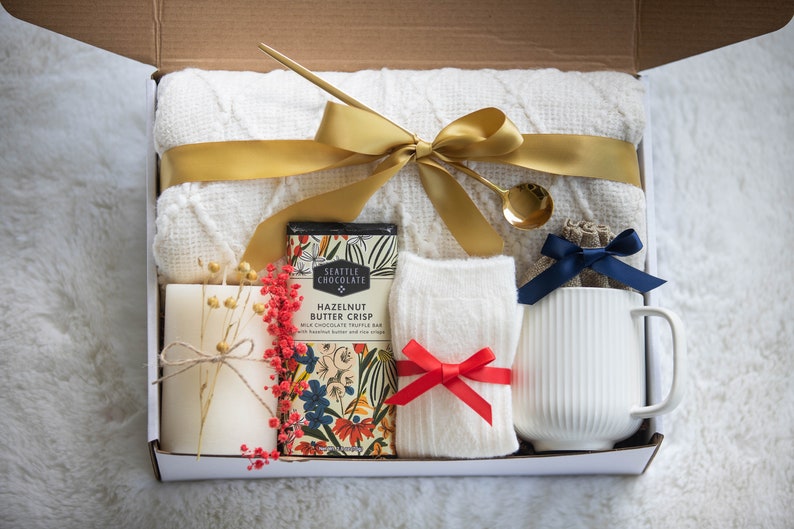 K&ouml;nnte beinhalten: Geschenkbox gef&uuml;llt mit einer wei&szlig;en Decke, einem goldenen Band, einer Schokoladenriegel, einer Kerze, Socken mit roter Schleife, einer Tasse und einem kleinen goldenen L&ouml;ffel. Die Box ist ge&ouml;ffnet.