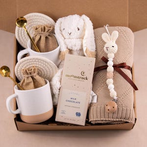 May include: A gift box filled with a white plush bunny, a tan knitted blanket, two white mugs with gold spoons, and a bar of milk chocolate. The mugs have a tan base and are paired with woven coasters and small burlap bags.