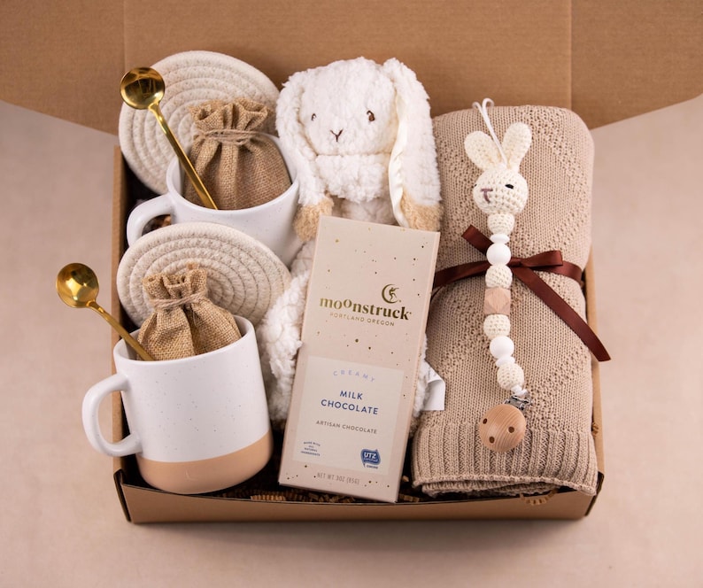 May include: A gift box filled with a white plush bunny, a tan knitted blanket, two white mugs with gold spoons, and a bar of milk chocolate. The mugs have a tan base and are paired with woven coasters and small burlap bags.
