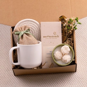 May include: A gift box filled with a white mug, a coaster, a small burlap bag tied with a green ribbon, a bar of chocolate, a gold spoon, and a small candle with three white rose-shaped candles. The box is lined with brown paper.