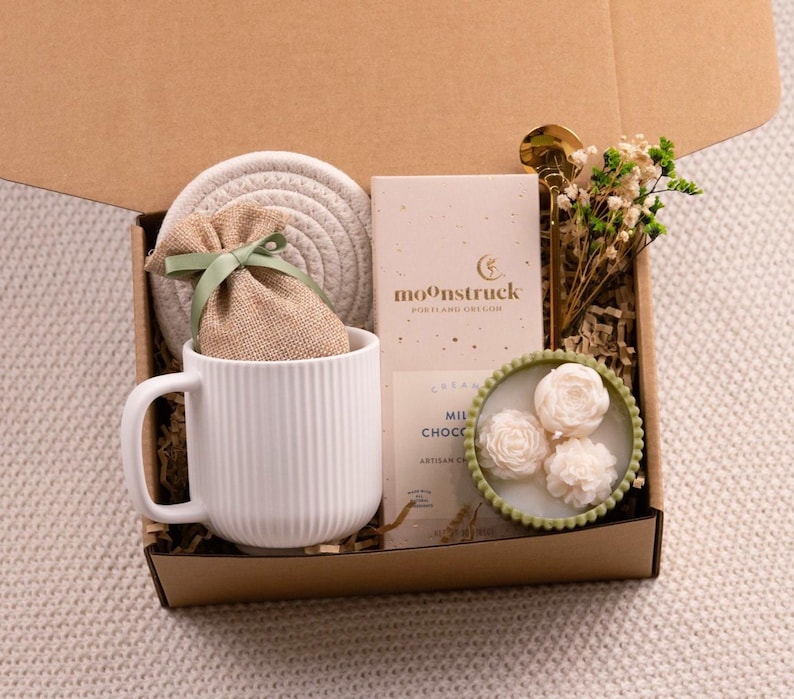 May include: A gift box filled with a white mug, a coaster, a small burlap bag tied with a green ribbon, a bar of chocolate, a gold spoon, and a small candle with three white rose-shaped candles. The box is lined with brown paper.
