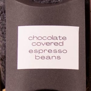 Puede incluir: Una caja negra en forma de almohada con una etiqueta blanca que dice "chocolate covered espresso beans". La caja está colocada sobre un fondo gris texturizado. El producto es probablemente un artículo de comida gourmet.