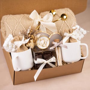 May include: A gift basket filled with a beige knit blanket, two white mugs, marshmallows, chocolates, and decorative items. The mugs have a vertical ribbed design. The basket is tied with white ribbons and bows.
