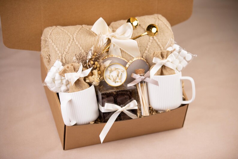 May include: A gift basket filled with a beige knit blanket, two white mugs, marshmallows, chocolates, and decorative items. The mugs have a vertical ribbed design. The basket is tied with white ribbons and bows.