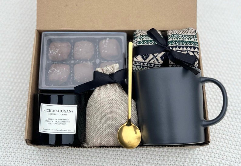 May include: A gift box filled with a black mug, a candle labelled "Rich Mahogany Scented Candle", a small bag of brown sugar, a gold spoon, and a pair of black and green patterned socks.