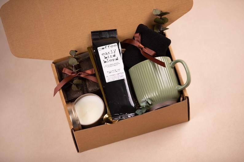 K&ouml;nnte beinhalten: Geschenkbox mit einer gr&uuml;nen Tasse, schwarzem Kaffee, einer Kerze und Schokolade. Die Tasse hat vertikale Rillen. Auf dem Kaffeebeutel steht "early bird blend". Die Box ist mit einem braunen Band gebunden.
