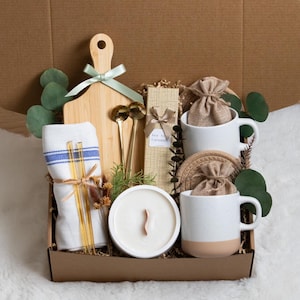 May include: A gift box filled with a wooden cutting board, two white mugs with tan bottoms, a candle, a towel, gold spoons, and other items. The box is brown and contains decorative greenery and a ribbon.