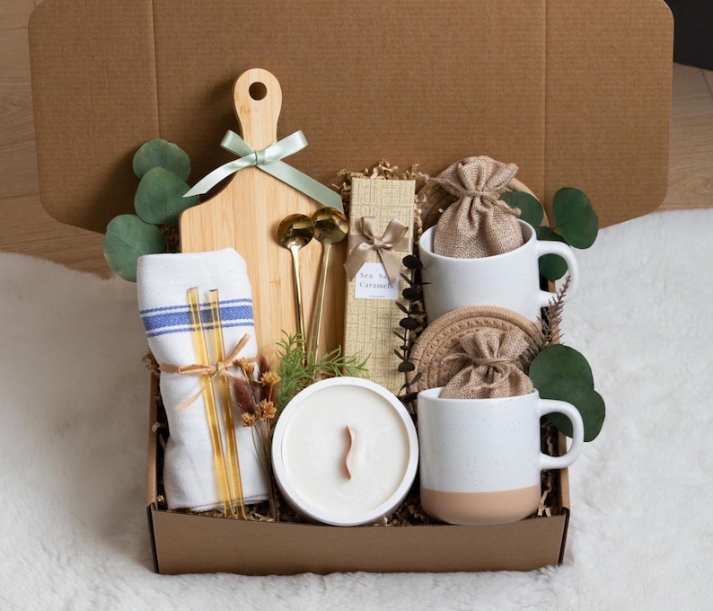 May include: A gift box filled with a wooden cutting board, two white mugs with tan bottoms, a candle, a towel, gold spoons, and other items. The box is brown and contains decorative greenery and a ribbon.