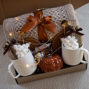 May include: A gift box containing a beige knit blanket with a rust-colored satin bow, two white ribbed ceramic mugs filled with white marshmallows, a small orange ceramic pumpkin, two burlap pouches, and string lights.
