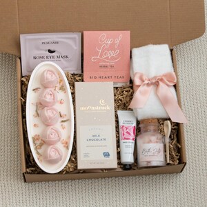 May include: A gift box filled with self-care items. Includes a rose eye mask, herbal tea, a white towel with a pink bow, rose-shaped soaps, milk chocolate, hand cream, and bath salts. The items are arranged in a brown cardboard box.