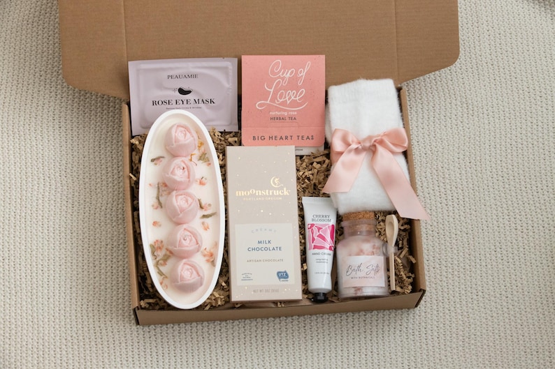 May include: A gift box filled with self-care items. Includes a rose eye mask, herbal tea, a white towel with a pink bow, rose-shaped soaps, milk chocolate, hand cream, and bath salts. The items are arranged in a brown cardboard box.