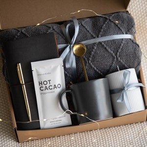 Peut inclure: Une bo&icirc;te en carton marron remplie d'un coffret cadeau douillet. Le coffret comprend une couverture en tricot gris, une tasse noire, une bougie blanche, un journal noir, un stylo dor&eacute; et un sachet de m&eacute;lange de chocolat chaud.