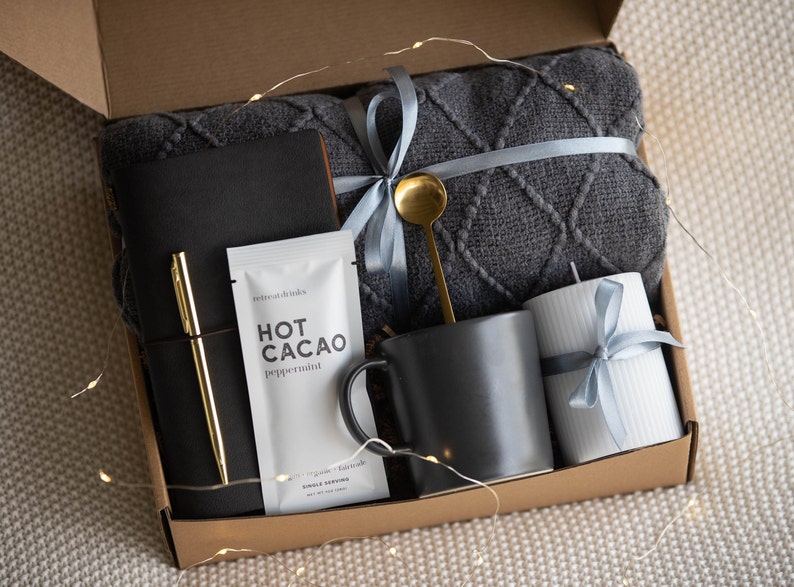 Peut inclure: Une bo&icirc;te en carton marron remplie d'un coffret cadeau douillet. Le coffret comprend une couverture en tricot gris, une tasse noire, une bougie blanche, un journal noir, un stylo dor&eacute; et un sachet de m&eacute;lange de chocolat chaud.