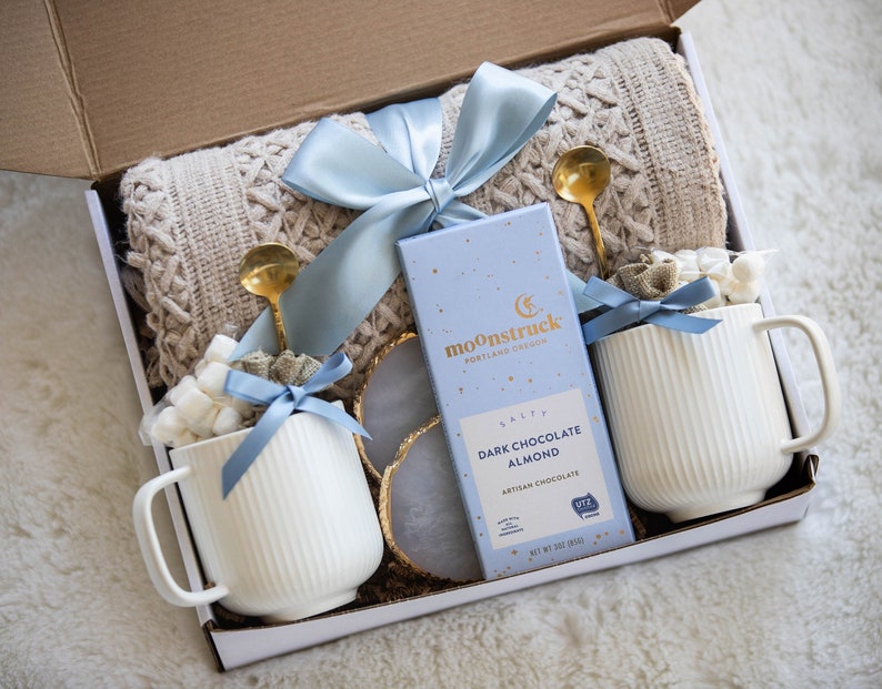 May include: A gift box with a cream-colored cable-knit blanket, two white ribbed mugs with marshmallows, gold spoons, a bar of dark chocolate, and a blue ribbon. The chocolate bar reads "Moonstruck Portland Design" and "Dark Chocolate Almond".