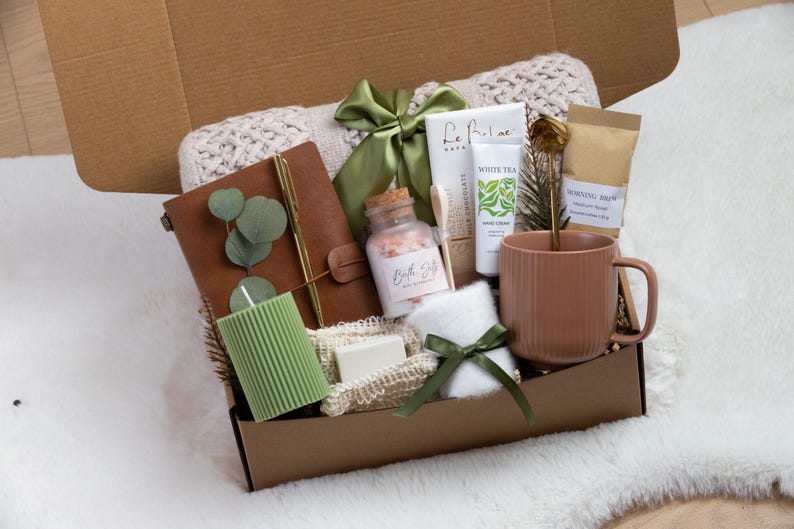 K&ouml;nnte beinhalten: Eine Geschenkbox mit Pflegeprodukten. Sie enth&auml;lt ein braunes Lederjournal, eine gr&uuml;ne Kerze, Badesalz, Handcreme, einen Becher und ein kleines Handtuch. Die Box ist mit einem gr&uuml;nen Band gebunden und enth&auml;lt eine wei&szlig;e Decke.