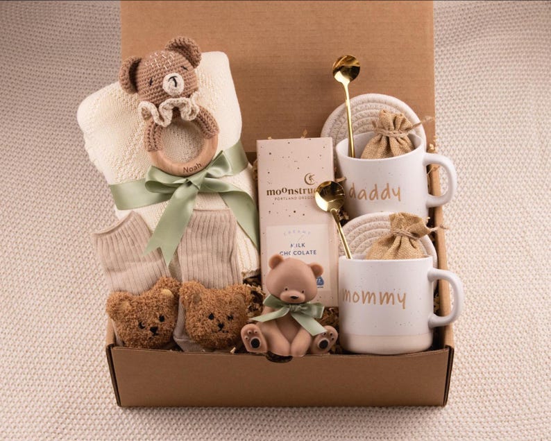 May include: A baby gift box containing a cream blanket, socks, and teddy bear toys. The box also includes two white mugs with "daddy" and "mommy" printed on them, chocolate, and gold spoons. A wooden baby rattle is also included in the gift set.