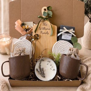 May include: A gift box filled with a variety of items. Includes two brown ribbed mugs, a wooden cutting board with text, a candle, coasters, and a box of dark chocolate sea salt caramels. The box is filled with decorative filler and greenery.
