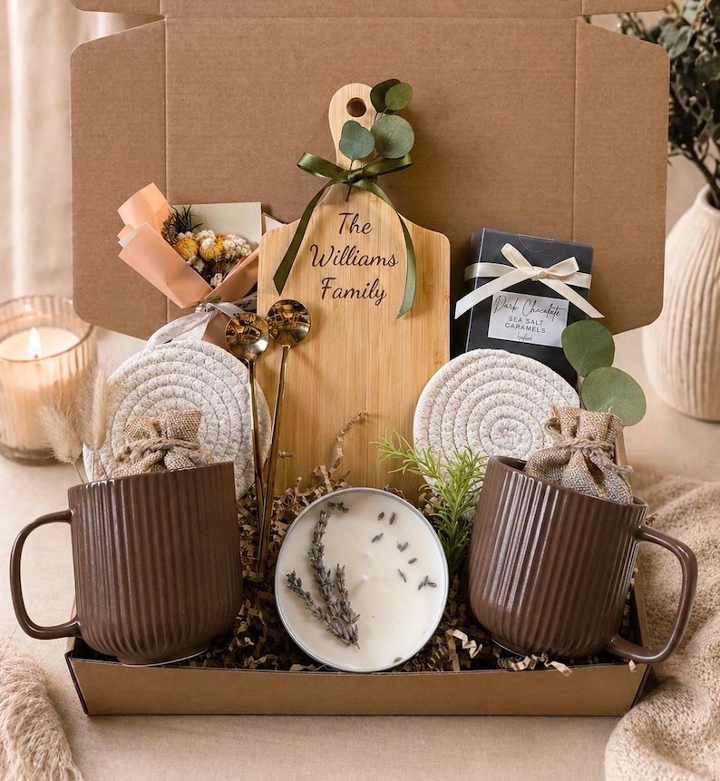 May include: A gift box filled with a variety of items. Includes two brown ribbed mugs, a wooden cutting board with text, a candle, coasters, and a box of dark chocolate sea salt caramels. The box is filled with decorative filler and greenery.