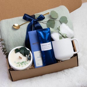 K&ouml;nnte beinhalten: Eine Geschenkbox gef&uuml;llt mit einer hellgr&uuml;nen Decke, einer wei&szlig;en Tasse, einer Kerze, dunkler Schokolade und Kaffee. Die Box ist mit einem marineblauen Band gebunden und enth&auml;lt einen goldenen L&ouml;ffel und Eukalyptuszweige. Die Artikel sind auf einer wei&szlig;en, flauschigen Oberfl&auml;che angeordnet.