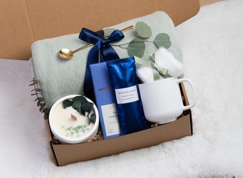 K&ouml;nnte beinhalten: Eine Geschenkbox gef&uuml;llt mit einer hellgr&uuml;nen Decke, einer wei&szlig;en Tasse, einer Kerze, dunkler Schokolade und Kaffee. Die Box ist mit einem marineblauen Band gebunden und enth&auml;lt einen goldenen L&ouml;ffel und Eukalyptuszweige. Die Artikel sind auf einer wei&szlig;en, flauschigen Oberfl&auml;che angeordnet.