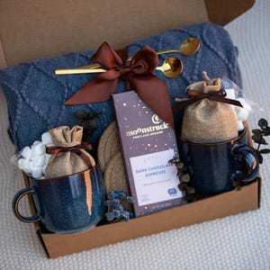 May include: A gift box containing a blue cable-knit blanket, two speckled blue ceramic mugs, two burlap pouches, a bar of Moonstruck Dark Chocolate Espresso, two gold-toned spoons, and two round woven coasters, all tied with a brown satin ribbon.