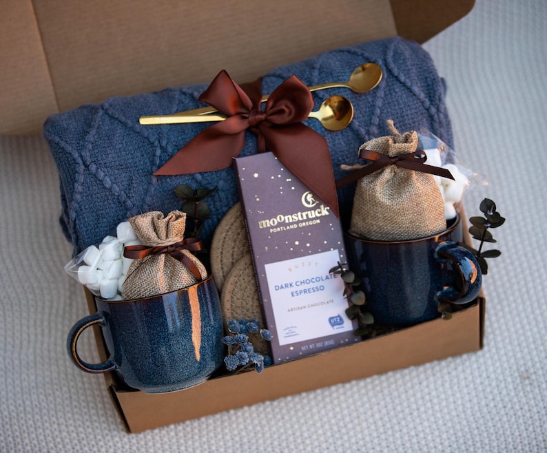 May include: A gift box containing a blue cable-knit blanket, two speckled blue ceramic mugs, two burlap pouches, a bar of Moonstruck Dark Chocolate Espresso, two gold-toned spoons, and two round woven coasters, all tied with a brown satin ribbon.