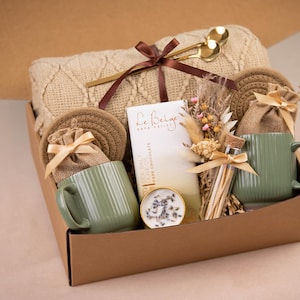 May include: A gift box filled with a beige knit blanket, two green mugs, coasters, and small burlap bags tied with ribbons. The box also contains a bar of dark chocolate, a small candle, and a bundle of dried flowers. Gold-colored spoons rest on top.