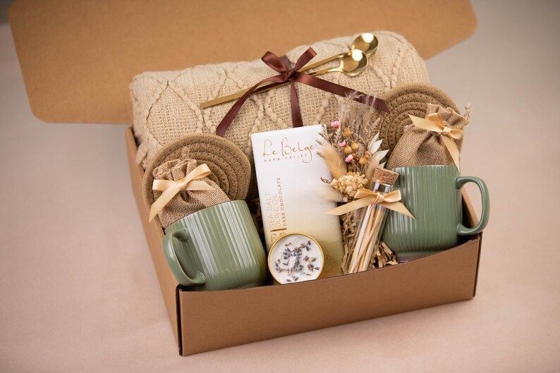 May include: A gift box filled with a beige knit blanket, two green mugs, coasters, and small burlap bags tied with ribbons. The box also contains a bar of dark chocolate, a small candle, and a bundle of dried flowers. Gold-colored spoons rest on top.