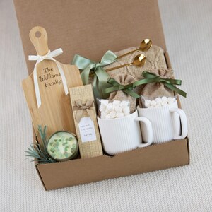 May include: A gift box containing a wooden paddle board with the text The Williams Family, a candle with green stones, a box of Sea Salt Caramels, two white ribbed mugs filled with marshmallows in burlap bags, and two gold spoons with a green ribbon.